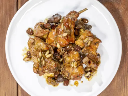 Pezzi di coniglio alla ligure, cotti con olive, pinoli e erbe aromatiche, serviti su un piatto bianco.