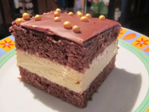 Pyszne milky way ciasto z czekoladowym blatem i kremowym nadzieniem, ozdobione złotymi kuleczkami.
