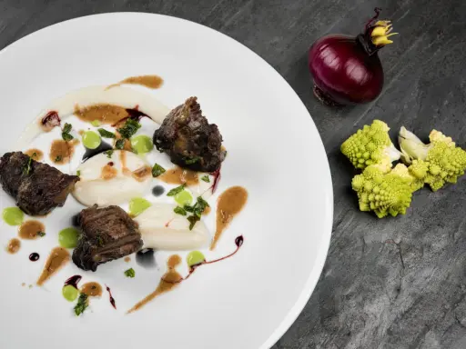 Succulenta guancia di manzo tenerissima, servita su purè bianco con gocce verdi e salsa bruna. Accanto, cipolla rossa e cavolfiore romanesco.