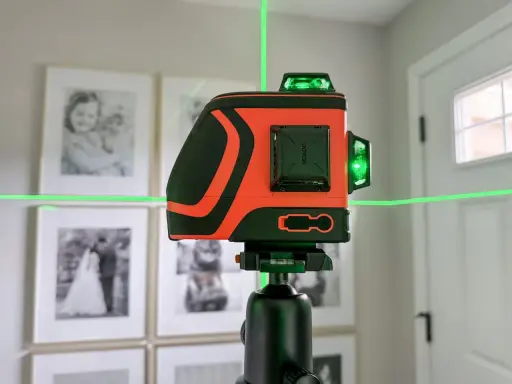 Pomarańczowa poziomica laserowa z zielonymi liniami na statywie, na tle oprawionych zdjęć.