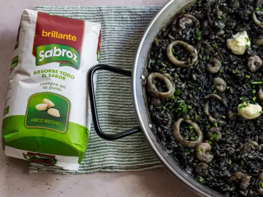 Arroz negro con calamares y gambas en paellera, junto a un paquete de arroz redondo Sabroz.