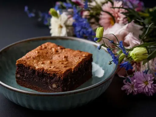 Un délicieux brownie healthy, parsemé de noix, dans un bol bleu, avec un bouquet de fleurs colorées en arrière-plan.