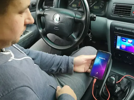 Mężczyzna w samochodzie pokazuje, jak połączyć telefon z autem za pomocą kabla i aplikacji Weblink. Na ekranie radia widoczne są ikony aplikacji.
