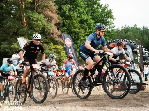 Kolarze biorą udział w wyścigu **zawody MTB**. Widać ich w akcji, jadących po piaszczystej trasie, z lasem w tle.