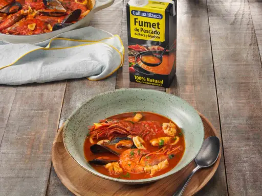 Un plato humeante de zarzuela de pescado, con gambas, mejillones y calamares, junto a un cartón de fumet de pescado.