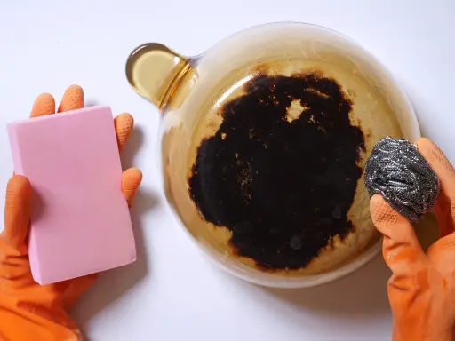 Manos con guantes naranjas sostienen una esponja rosa y un estropajo metálico, listos para aprender como quitar el quemado de una olla.