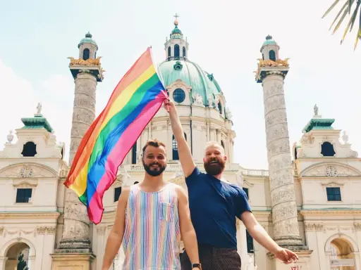 Two men proudly hold a rainbow flag in front of a grand building in Austria, celebrating gay pride.