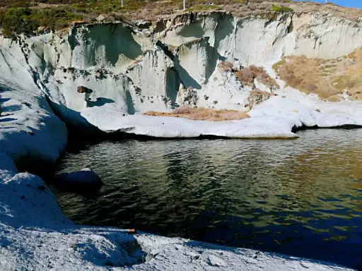 Scogliere bianche erose dal mare, con vegetazione secca e un'acqua scura che riflette il cielo. Sembra un luogo selvaggio, quasi un "cane malu" che custodisce la costa.