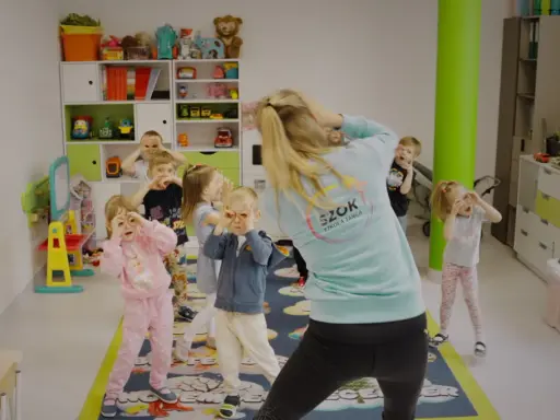 Dzieci na zajęciach tańca wykonują proste układy taneczne do znanych piosenek, naśladując ruchy nauczycielki.