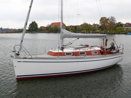 Ein Mann steuert die Segelyacht BIGA 242 auf ruhigem Wasser, im Hintergrund Häuser und Bäume.