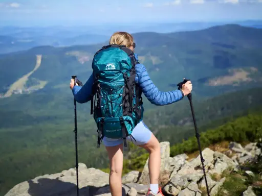 Kobieta z turkusowym plecakiem w góry, z kijkami trekkingowymi, podziwia panoramę.