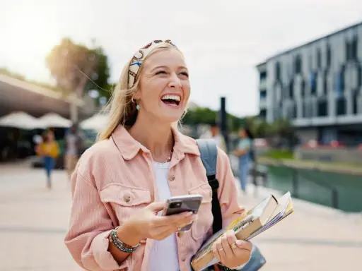 Szczęśliwa studentka z polską maturą, która planuje studia za granicą, śmieje się, trzymając telefon i książki.