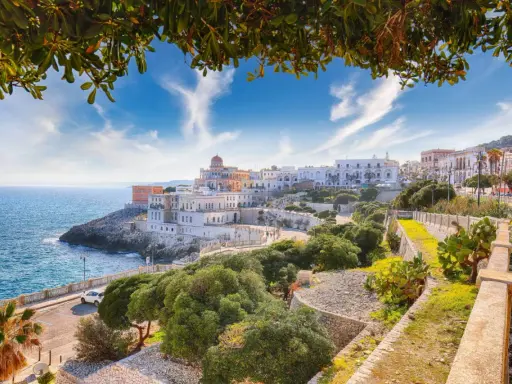 Panoramica di un borgo costiero con edifici bianchi e colorati, mare blu e vegetazione mediterranea. Ecco cosa vedere a La Maddalena, un gioiello pugliese.