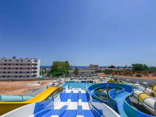 Kolorowe zjeżdżalnie wodne i baseny w Narcissos Waterpark Resort, z hotelami i morzem w tle.