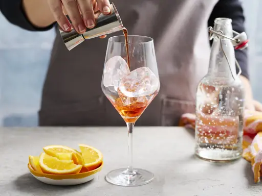 Ein Aperol-Spritz-Cocktail mit Prosecco wird zubereitet. Eiswürfel im Glas, daneben Orangenstücke und eine Flasche Sprudelwasser.