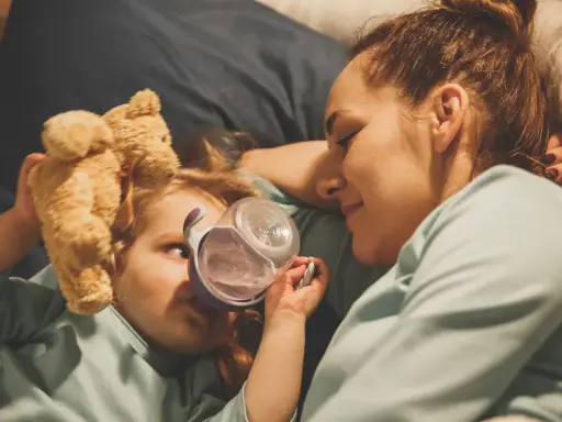 Mama opiekuje się dzieckiem z rotawirusem. Podaje mu wodę z kubeczka, a obok leży pluszowy miś.