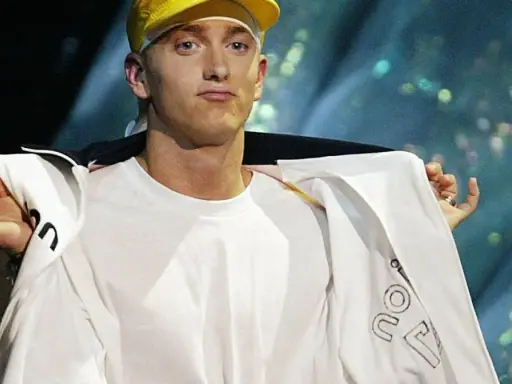 Slim Shady w żółtej czapce i białej bluzie, z gestem dłoni.