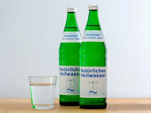 Zwei grüne Flaschen Heilwasser und ein Glas Wasser. Ein Glas Wasser vor dem Schlafengehen trinken kann beim Abnehmen helfen.