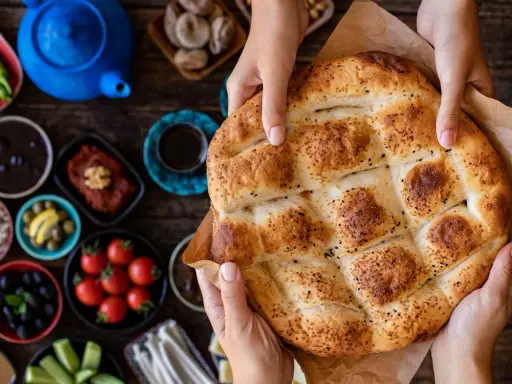 Hände halten ein frisch gebackenes Ramadan Brot, bestreut mit Sesam und Schwarzkümmel. Daneben liegen kleine Schalen mit Oliven, Tomaten und anderen Köstlichkeiten.