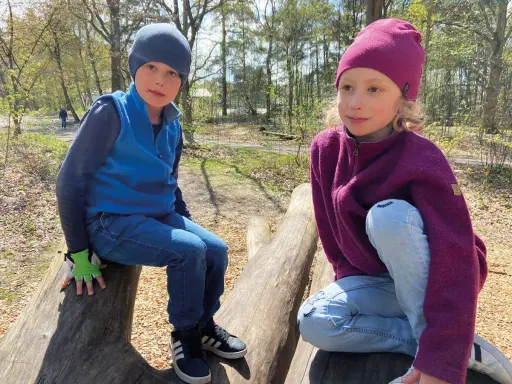 Två barn sitter på stockar i skogen. Båda bär en mjuk merinoull mössa barn, perfekt för utomhuslek.