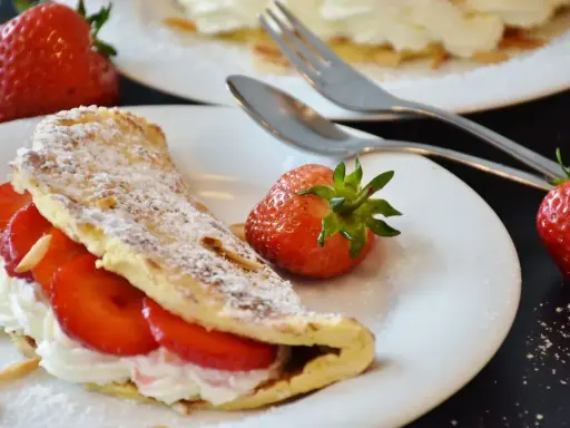 Puszysty omlet z białek, nadziany kremem i truskawkami, posypany cukrem pudrem. Obok leżą świeże owoce i sztućce.
