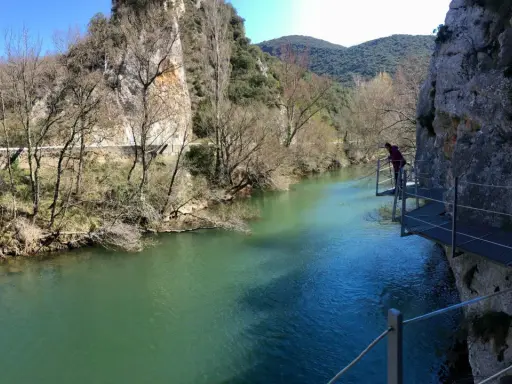 Persona camina por pasarela metálica junto a río turquesa, explorando las rutas de Burgos. Paisaje rocoso y vegetación de ribera.