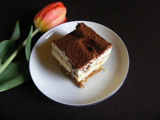 Pyszny kawałek tiramisu, jednego z 10 ciast bez pieczenia, obok czerwonego tulipana.