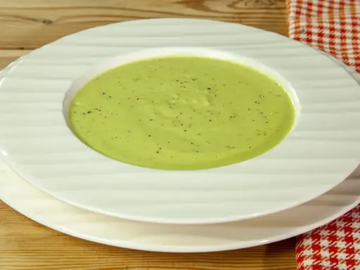 Una ciotola di cremosa crema di zucchine, condita con pepe nero, servita su piatti bianchi su un tavolo di legno.