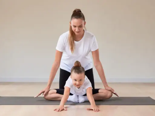 Frau und Kind machen Yoga. Das Kind krabbelt auf der Matte, die Frau unterstützt es. Yoga mit Kindern unter 3 Jahren fördert spielerisch die Bewegung.