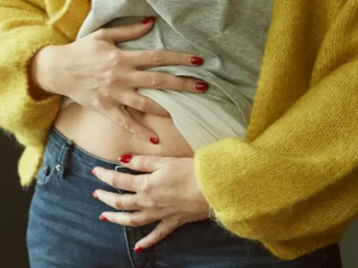 En kvinna med röda naglar håller om sin mage, klädd i en gul kofta och jeans. Hon känner sig gravid trots mens.