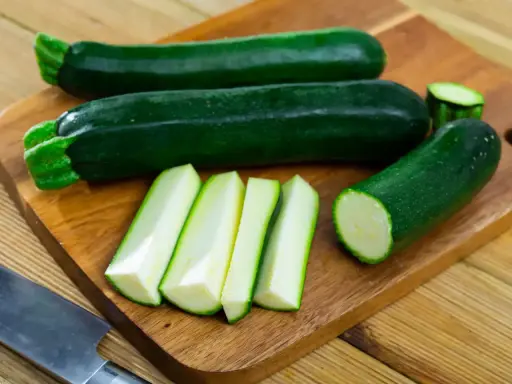Courgettes vertes entières et coupées en bâtonnets sur une planche à découper, prêtes à congeler.