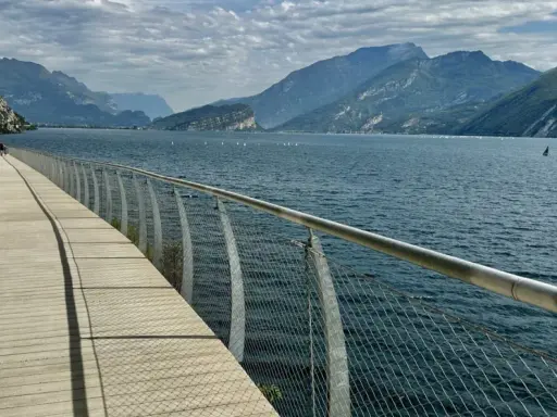 Ścieżka spacerowa nad jeziorem Garda, z widokiem na góry i żaglówki. Idealne miejsce, by zobaczyć piękno tego regionu.