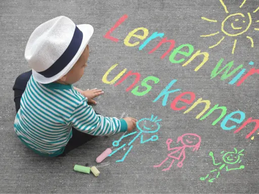 Kleiner Junge mit Hut malt mit Kreide auf Asphalt. Namensspiele für Kinder: "Lernen wir uns kennen!" mit Sonne und Figuren.