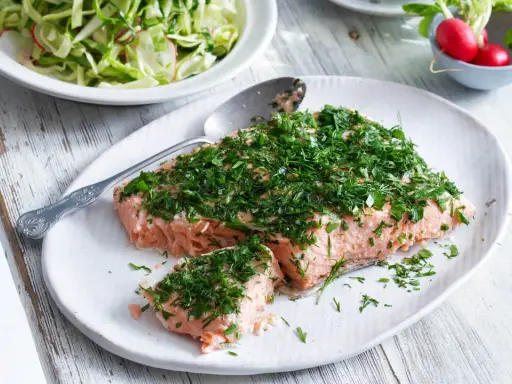 En saftig bit confiterad lax, generöst toppad med färska örter, serveras på en vit tallrik. Bredvid syns en fräsch sallad och några röda rädisor.