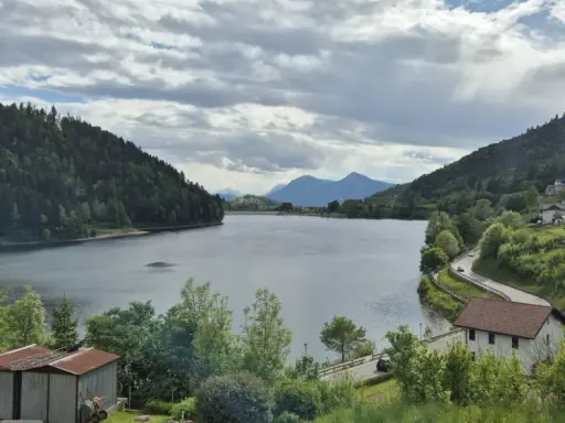 Panoramica del lago di Stramentizzo circondato da montagne verdi e un villaggio pittoresco. Nuvole minacciose nel cielo.