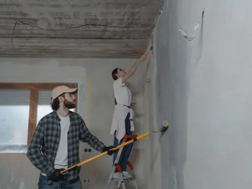 Para z zaangażowaniem wykonuje poprawki na pomalowanej ścianie. On trzyma wałek, ona stoi na drabinie.