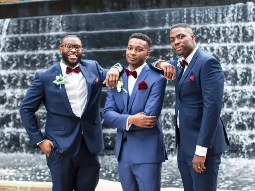 Three groomsmen in stylish navy blue suit combinations, featuring burgundy bow ties and pocket squares, pose in front of a waterfall.