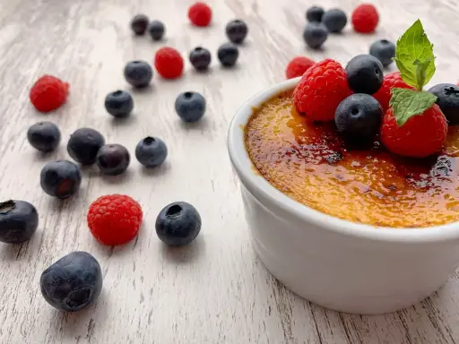 Creme brulee mit karamellisierter Kruste, garniert mit frischen Himbeeren und Blaubeeren, umgeben von weiteren Beeren auf hellem Holz.