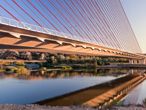 El puente de Castilla-La Mancha se refleja en el río, con sus cables rojos y el cielo azul.