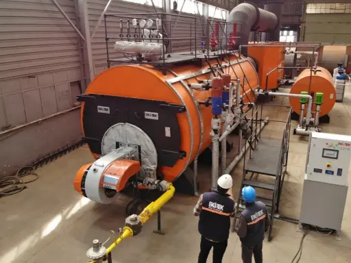 Dos técnicos observan varios tipos de calderas industriales naranjas y grises en una fábrica.