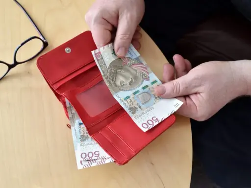 Ręce wkładają banknot 500 zł do czerwonego portfela. Obok okulary. Może to być pomocne przy sprawdzaniu 14 emerytury tabeli netto.