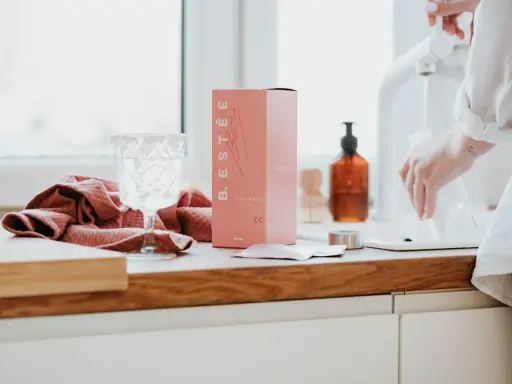 Różowe pudełko z kolagenem B. ESTÉE, szklanka wody i ręcznik. To estetyczne zdjecia produktowe.