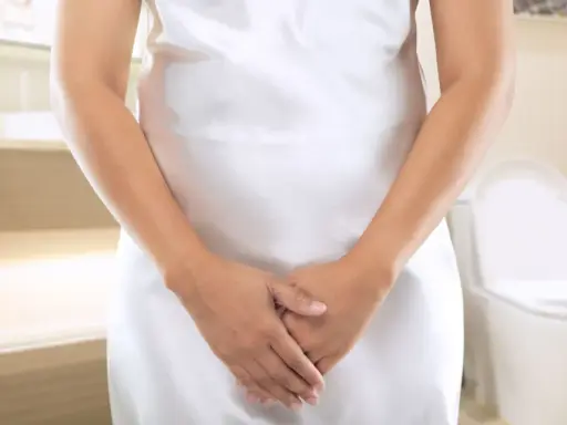 Kobieta w białej sukience w łazience, z dłońmi skrzyżowanymi na brzuchu. W tle toaleta. Szuka domowych sposobów na nietrzymanie moczu.