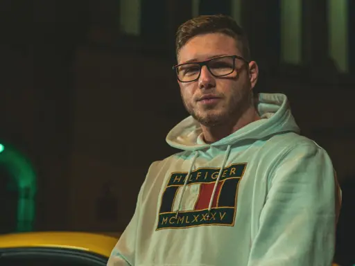 Kubańczyk cały ja utwory. Młody mężczyzna w okularach i bluzie z kapturem Tommy Hilfiger, nocą, obok żółtego samochodu.