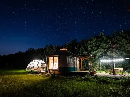 Nocne niebo nad Jurą Krakowsko-Częstochowską. Dwa noclegowe domki, jurt i kopuła, oświetlone ciepłym światłem, otoczone zielenią.