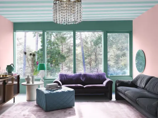 En färgstark salong med 30-tals inredning. Mörklila soffa, grå soffa, turkos pall och rosa matta. Fönster med utsikt över skog.