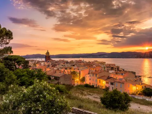 Sonnenuntergang über St. Tropez. Die Sonne versinkt im Meer, beleuchtet die Stadt und den Kirchturm in warmen Farben.