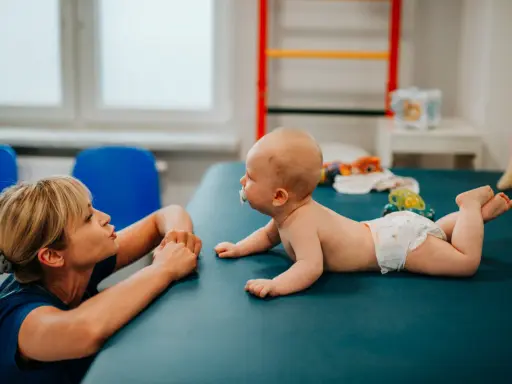 Terapeuta zachęca 3 miesięczne dziecko, które nie chce leżeć na brzuchu, do ćwiczeń na macie.