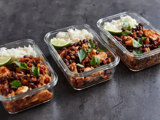 Meal prep für Muskelaufbau: Drei Glasbehälter mit Reis, schwarzen Bohnen, Hähnchen und Limette. Perfekte meal prep muskelaufbau rezepte für deine Fitnessziele.