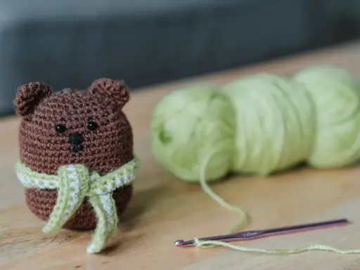 Os **sonajeros de crochet patrones gratis** son adorables. Un osito marrón con bufanda verde y blanca, junto a ovillos de lana y una aguja de crochet.